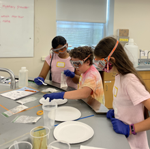 Three day camp participants standing at a bench in a lab participate in a chemistry activity. 
