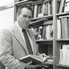 Black and white photo of John Hummel standing next to a book shelf holding an open book in his hands.