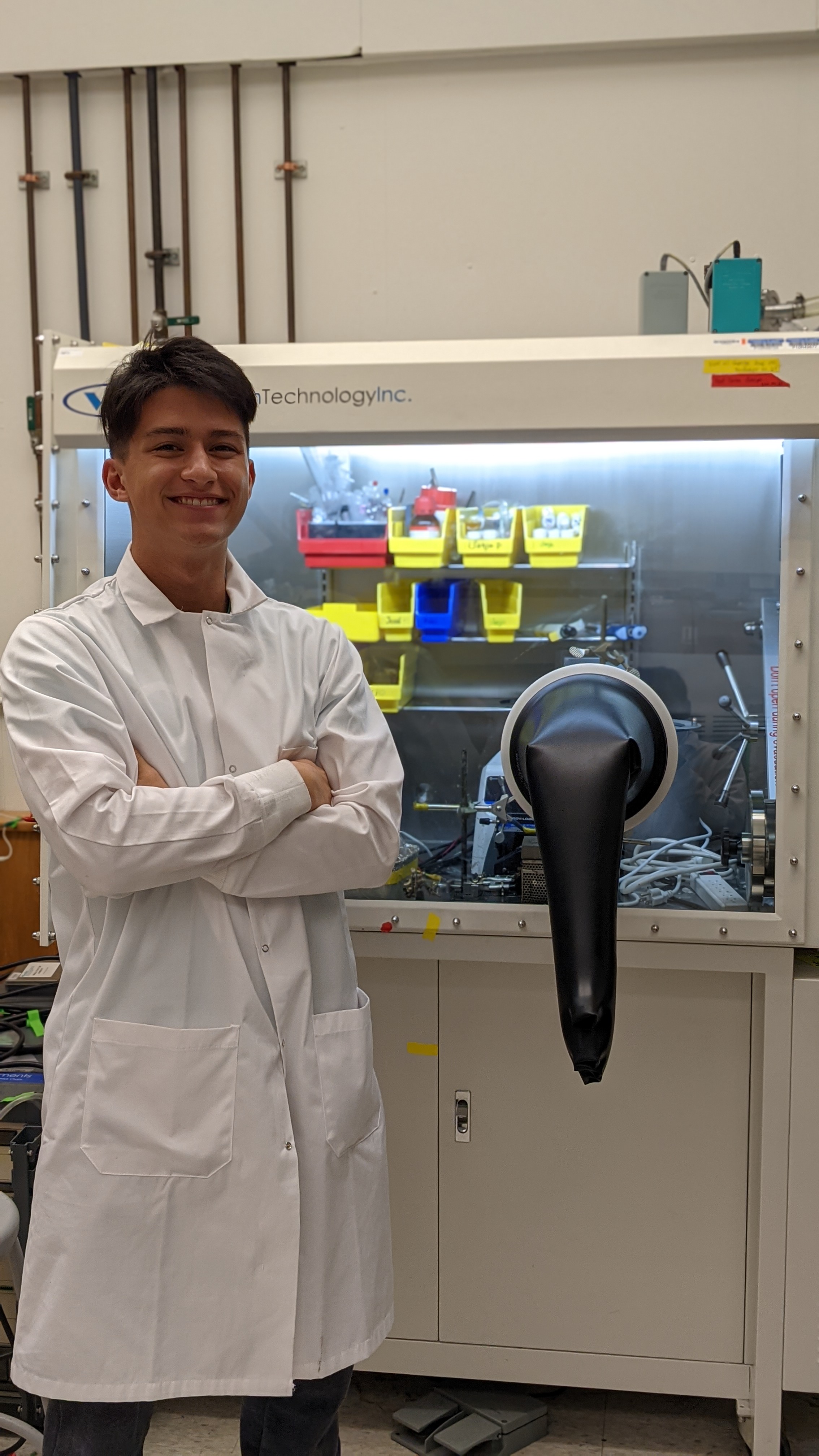 Curt Althaus wearing a white lab coat with arms folded standing in a lab in front of chemistry lab equipment.