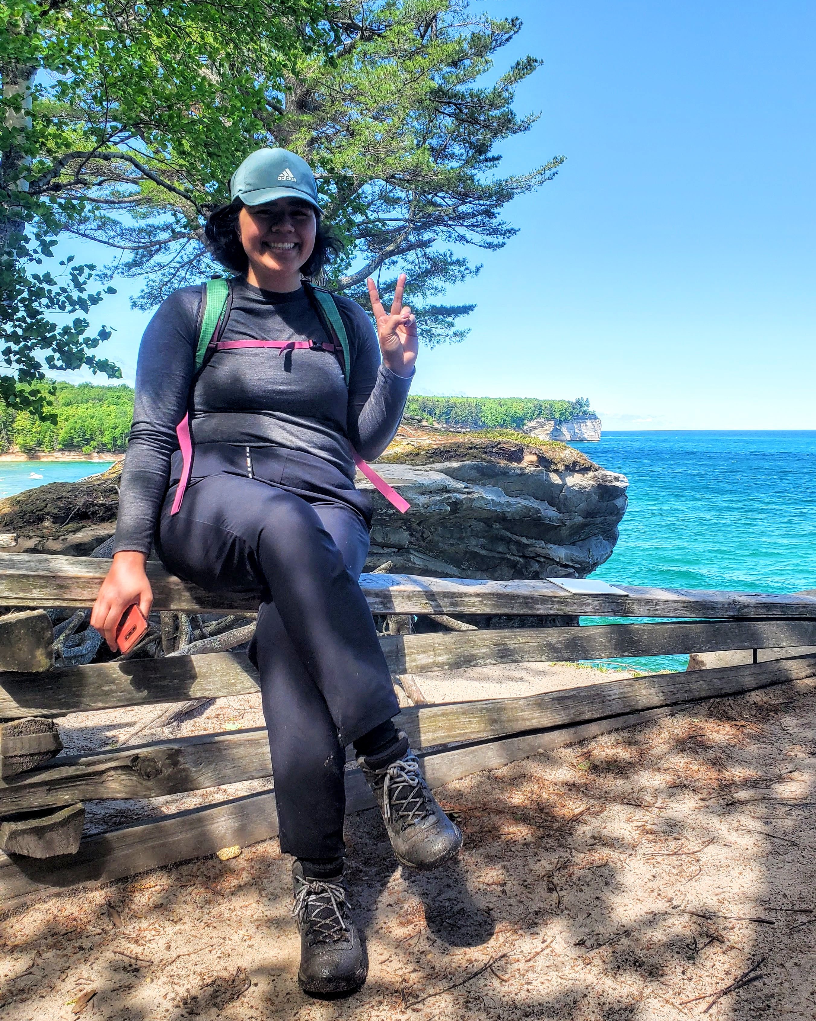 Tepora Su'a sits on a rock wall on a hiking trip with Lake Superior and trees in the background.