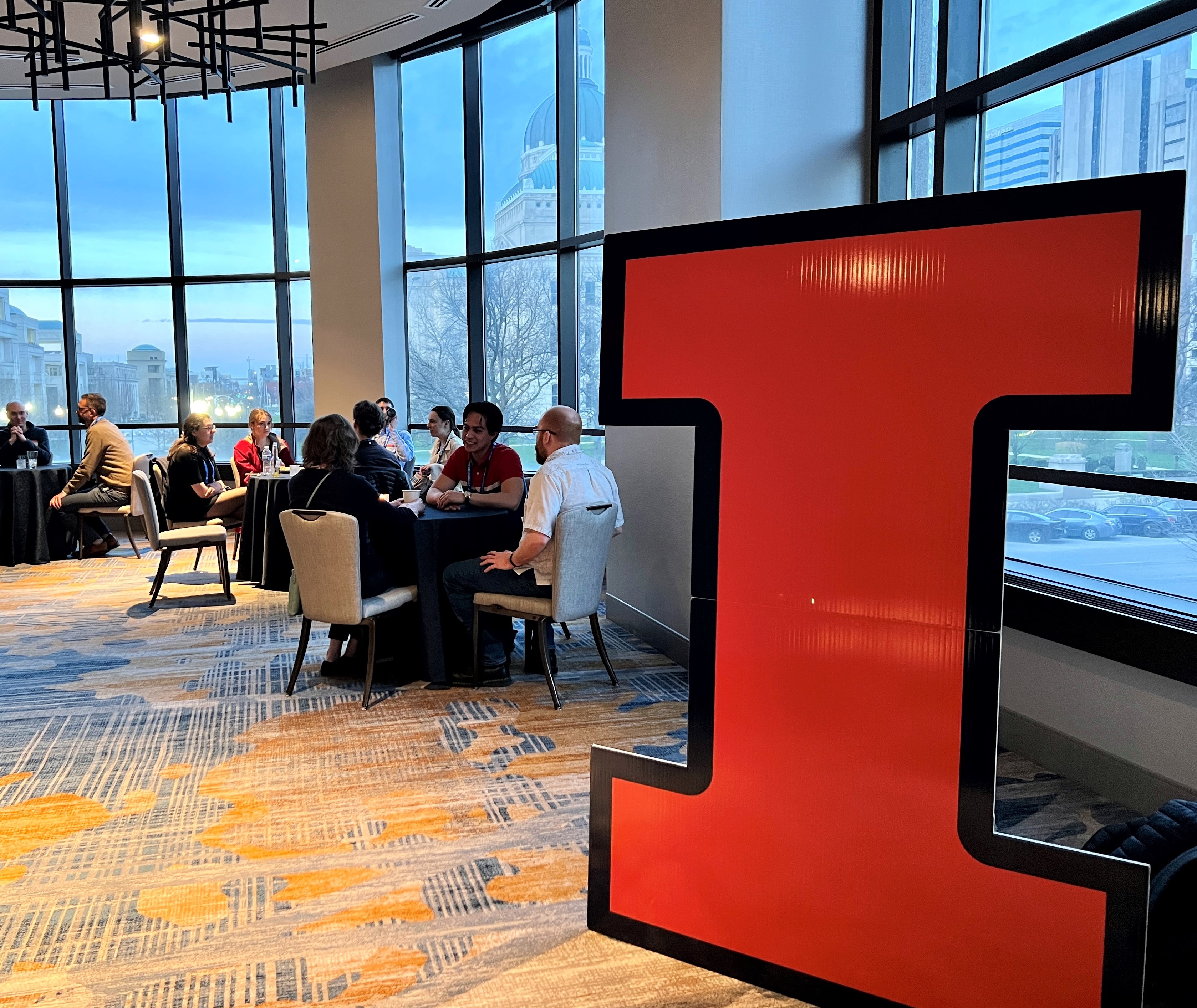 A large cardboard block I stands in the foreground of a photo of guests sitting at tables in the background at the reception location.