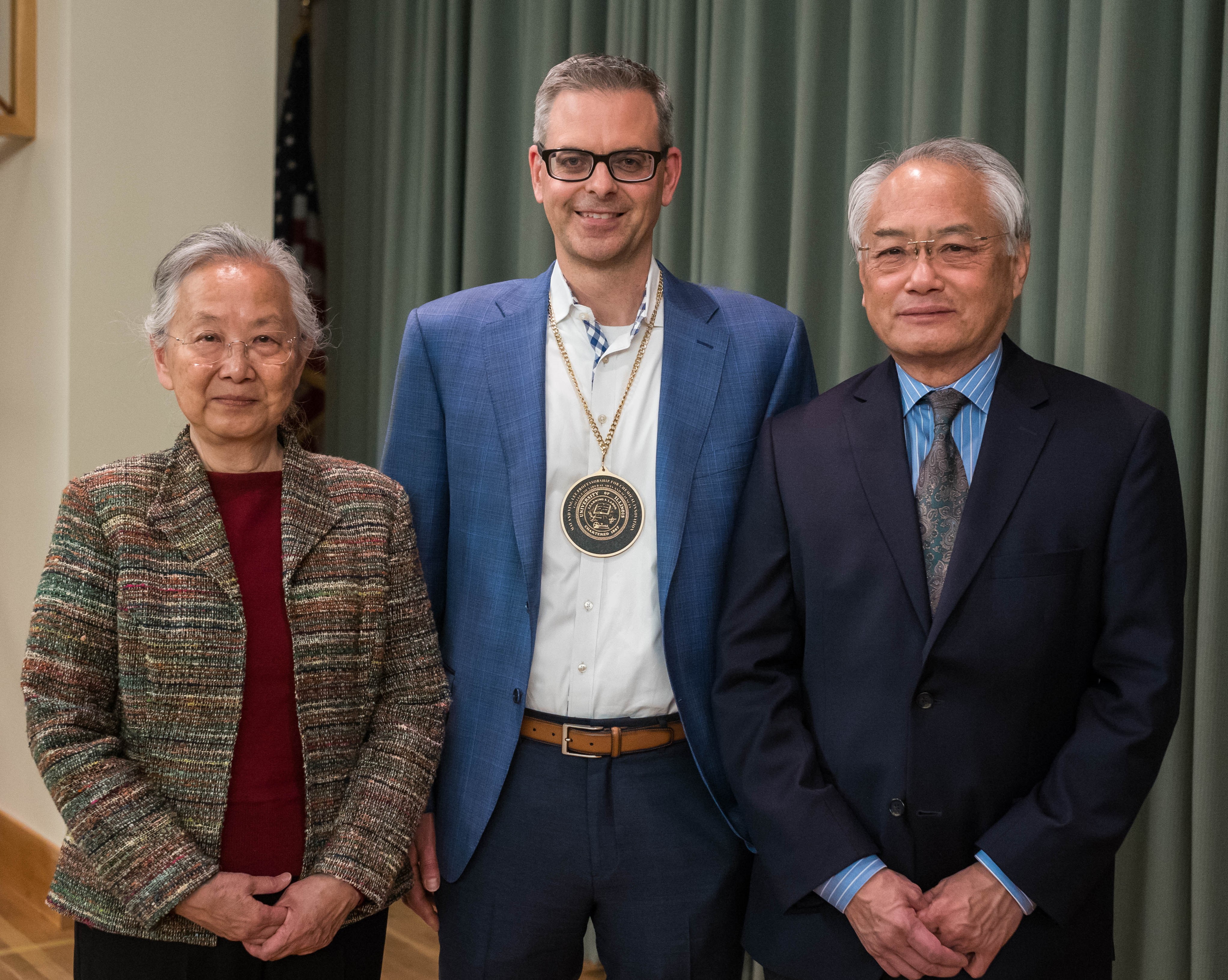 May and Ving Lee stand on either side of Martin Burke on a stage with a curtain behind them.