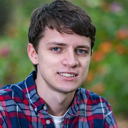 Outdoors portrait of Aidan Lindsay from the shoulders up and green foliage in the background.