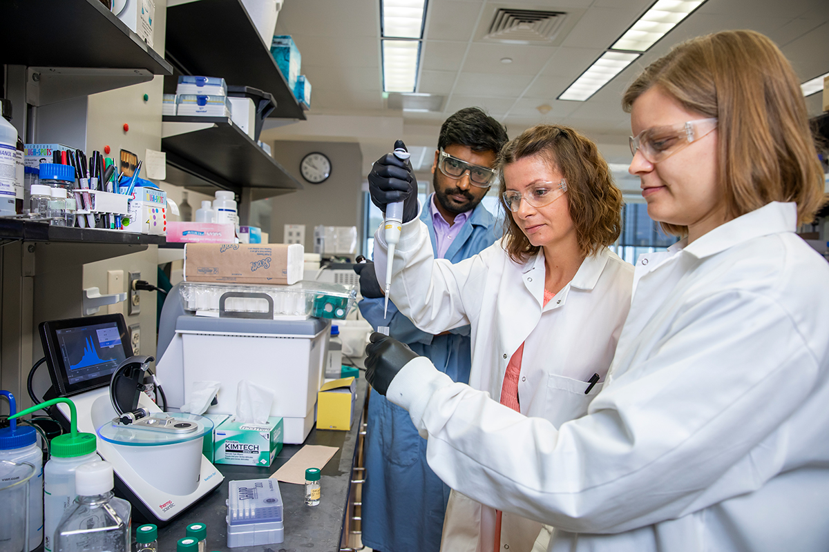 Three researchers in lab coats work at a bench in a lab on the UIUC campus.