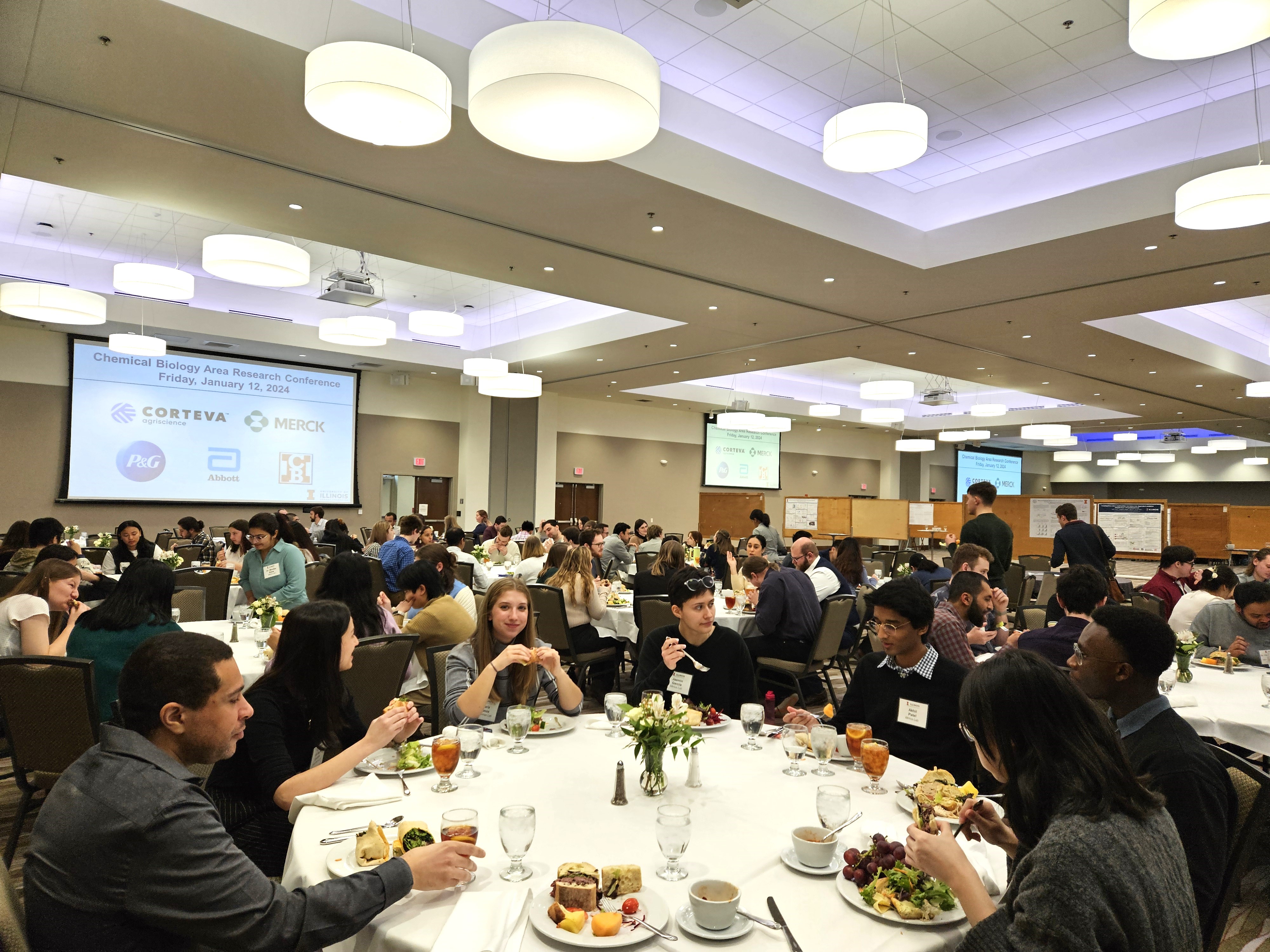 A large room full of round tables filled with people sitting at each table eating lunch.