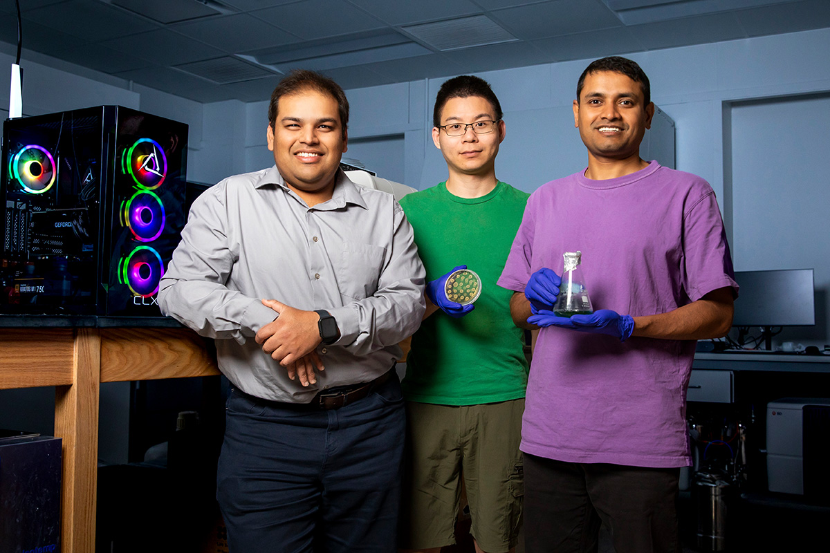 Angad Mehta and two other researchers stand side by side in one of their lab spaces with equipment behind them and holding equipment used in their study. 