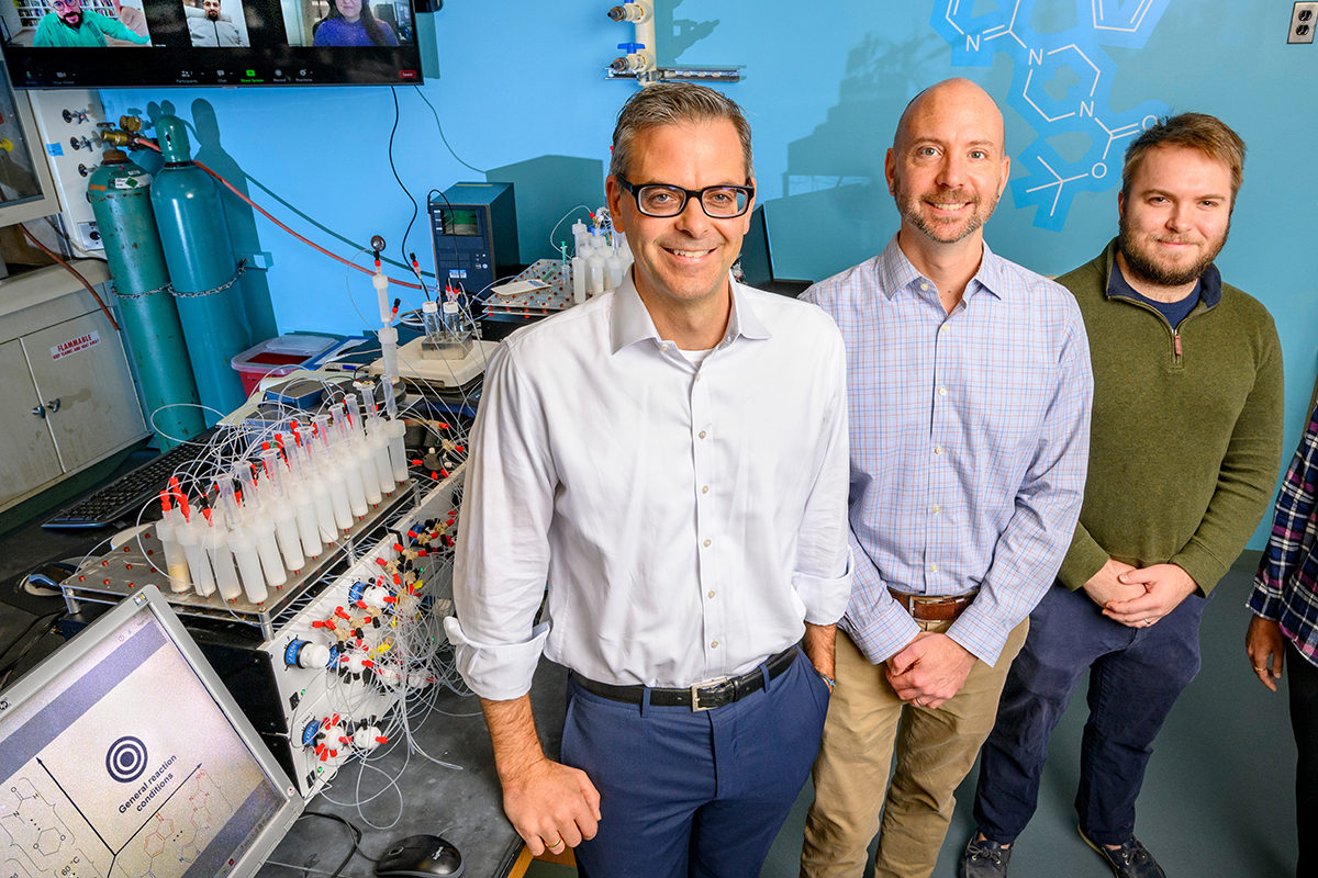 Three researchers stand side by side near equipment in a chemistry lab