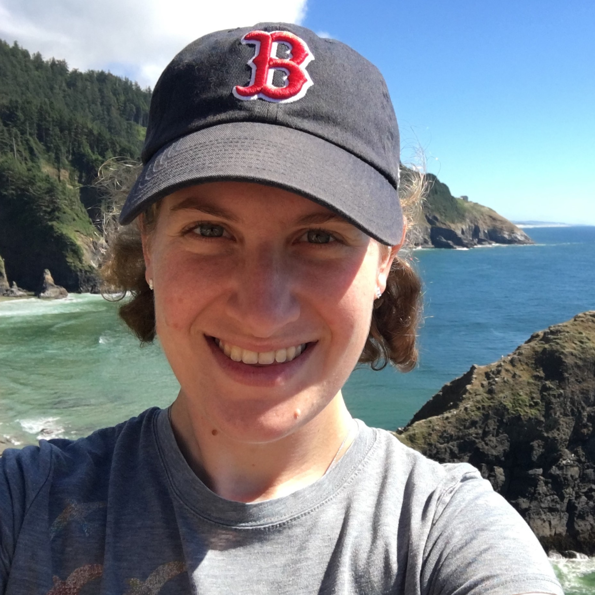 Head shot of Sarah Perlmutter in a baseball cap standing in front of an ocean view