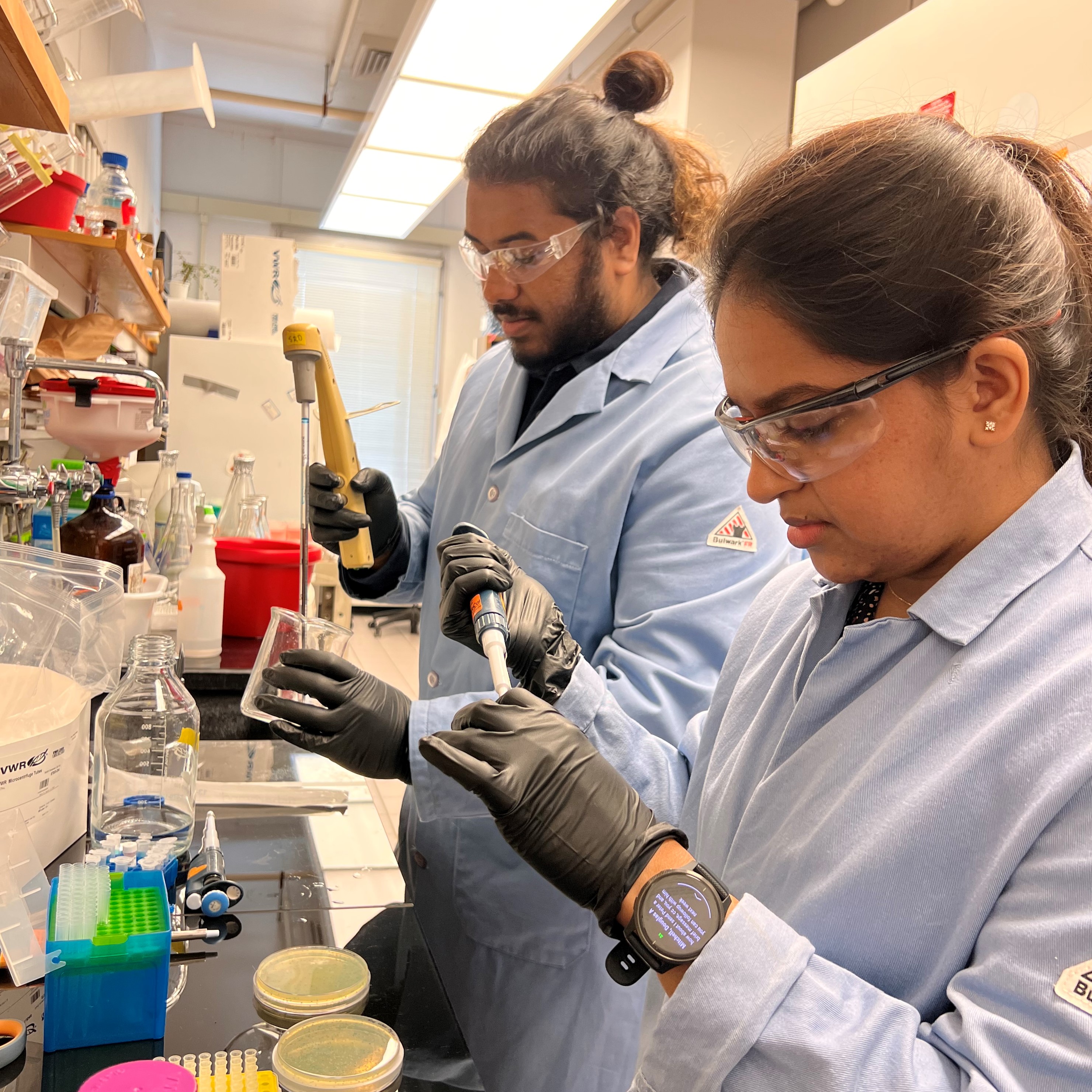 Two students standing side by side working in the lab in lab coats and PPE