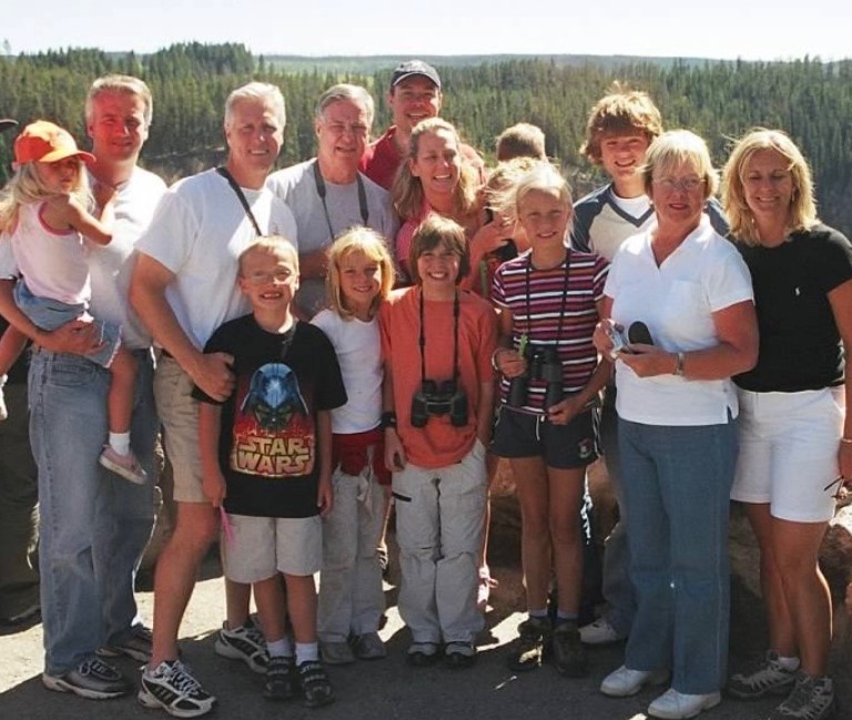 Dr. Milligan and 13 of his family members stand in a group in a location at Yellowstone overlooking a wooded area.