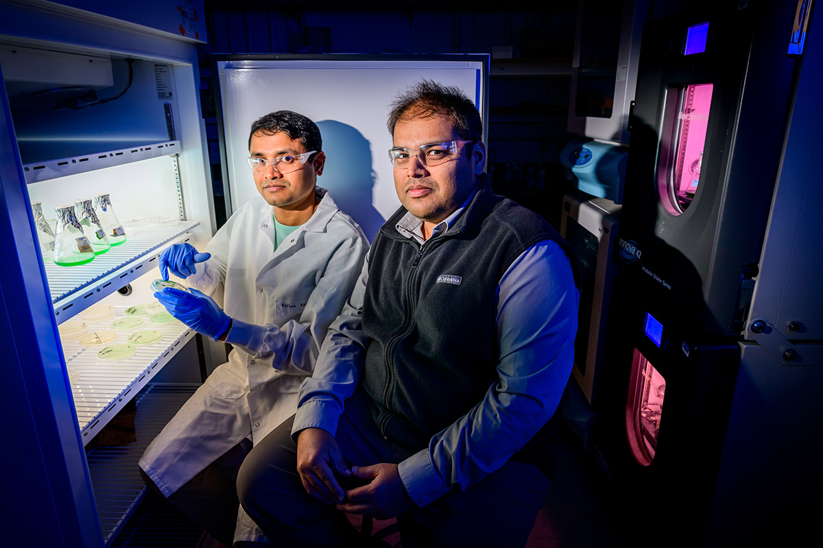 Two researchers in a dark lab sit in front of a refrigerator that stores research items. 