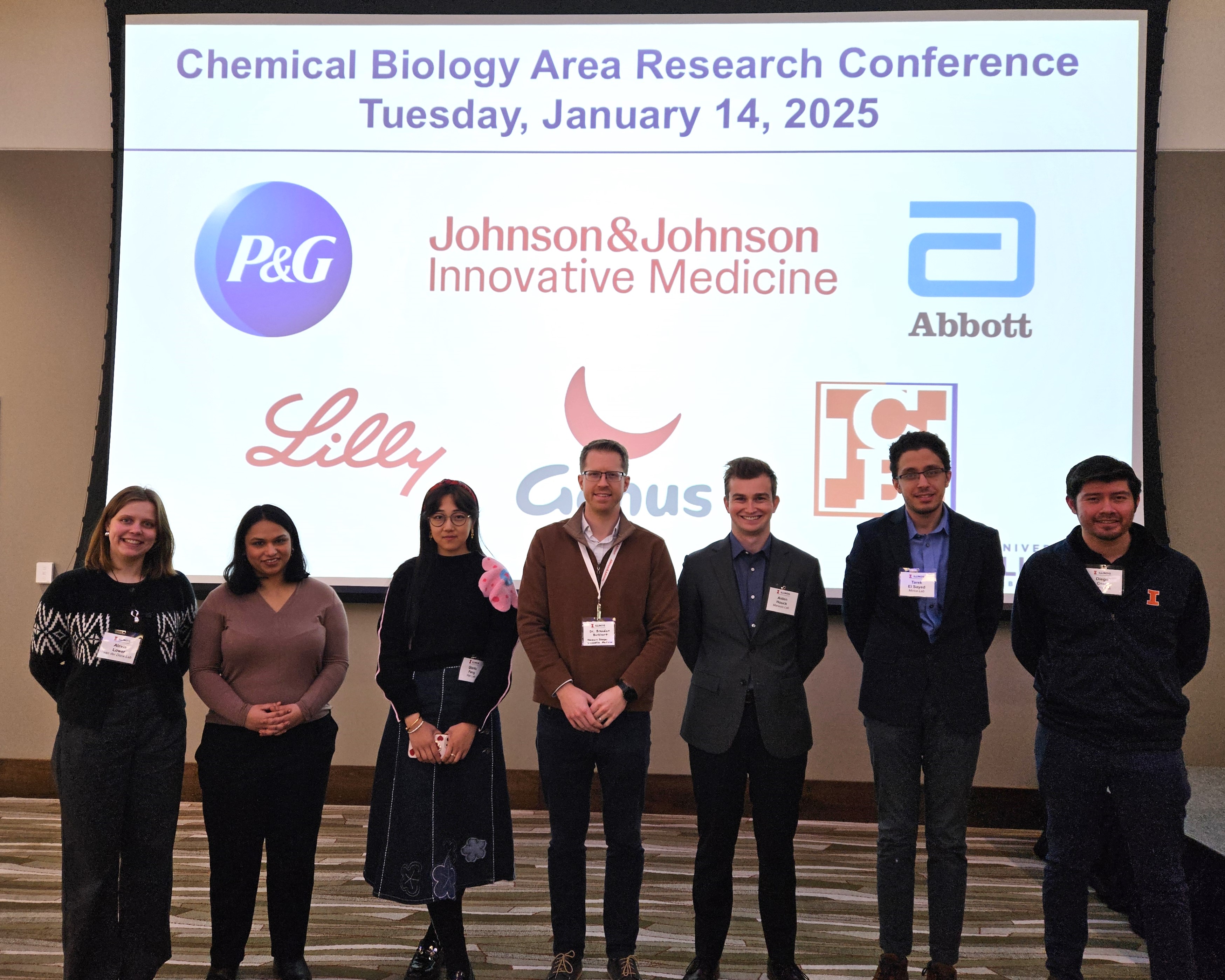 Group of graduate student presenters standing side by side in front of a large screen with conference sponsor logos. 