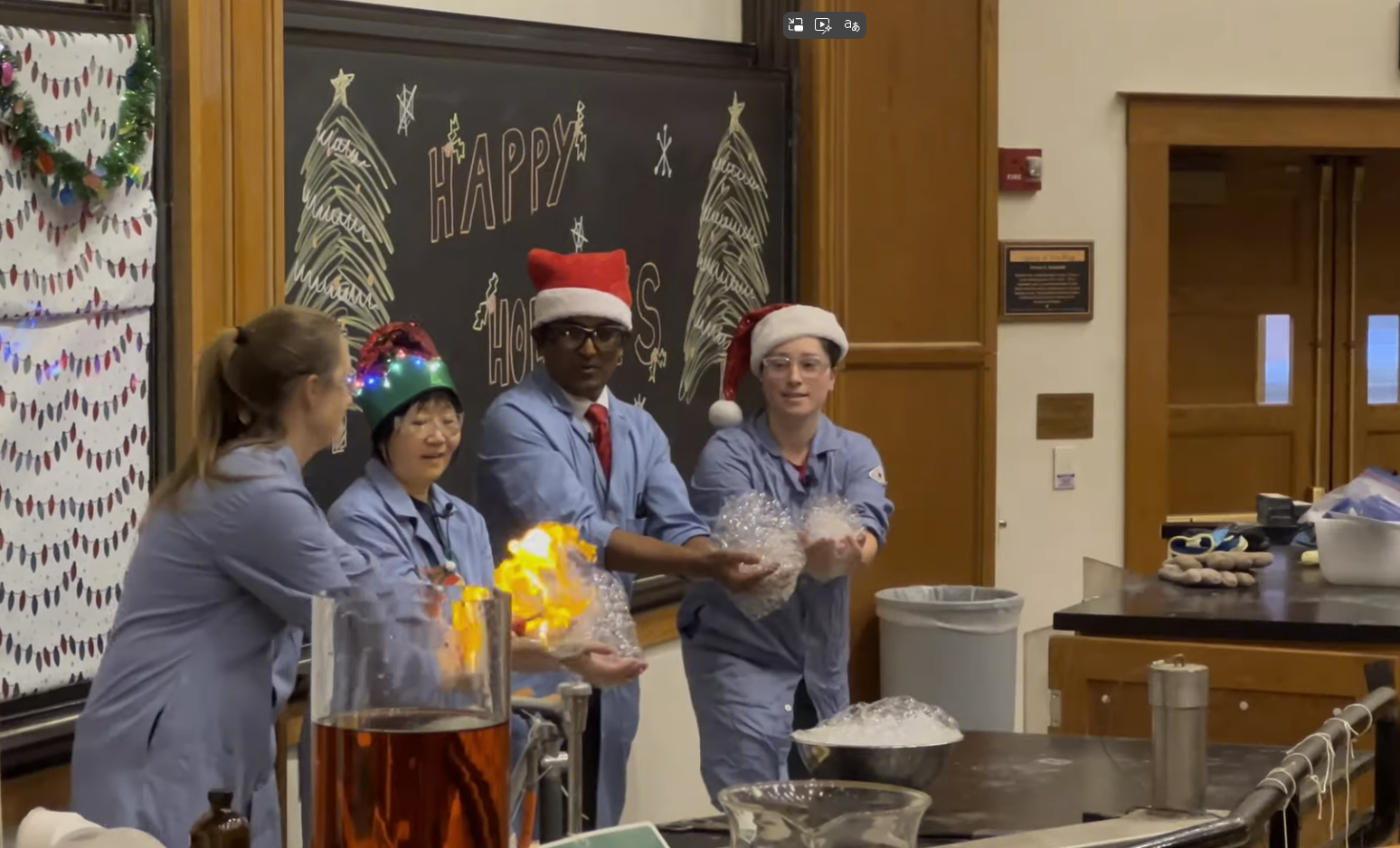 Four people in lab coats and holiday hats doing a chemistry demonstration with fire in a classroom