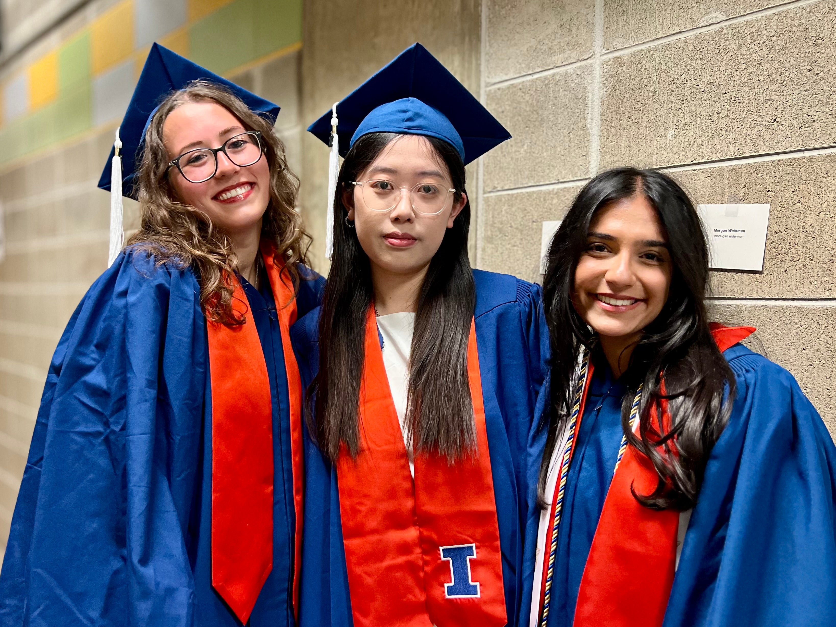 Three chemistry graduates in blue caps and gowns standing together in front of a cement wall