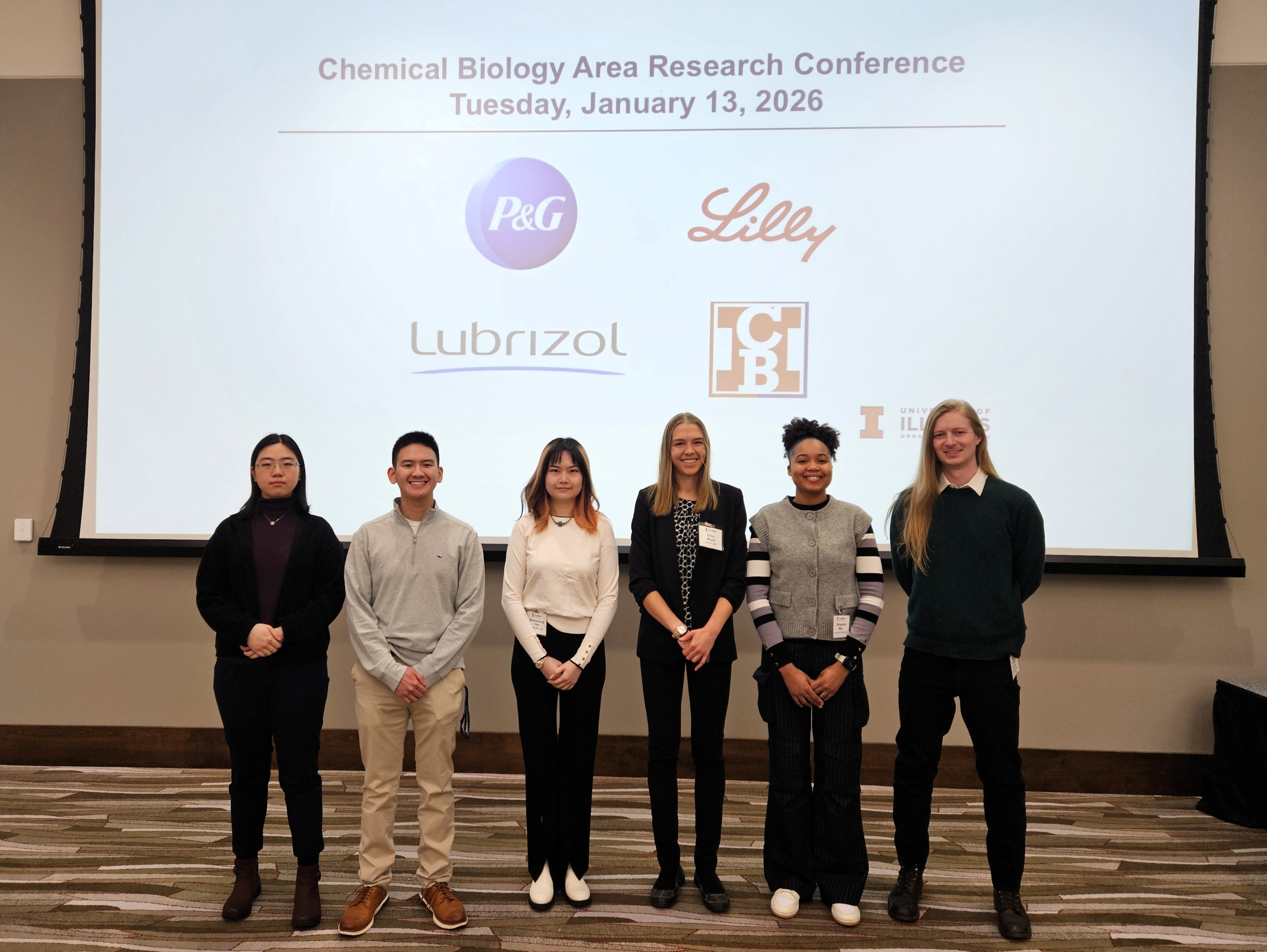 Six graduate student presenters standing side by side in front of a large overhead screen with names of conference sponsors.