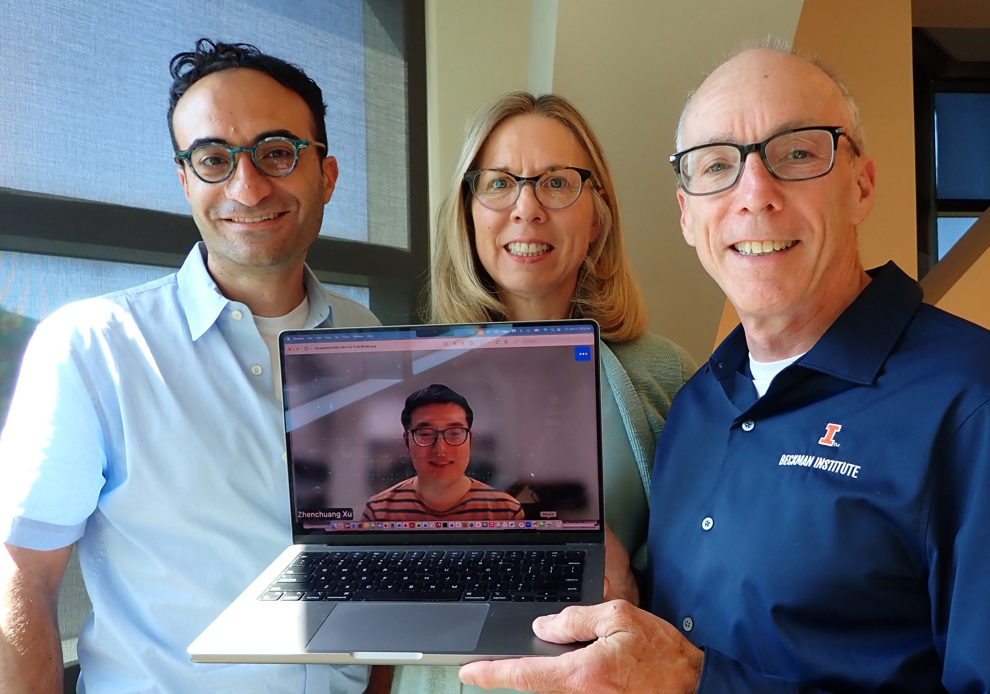 Three people standing side by side holding a laptop with image of a person