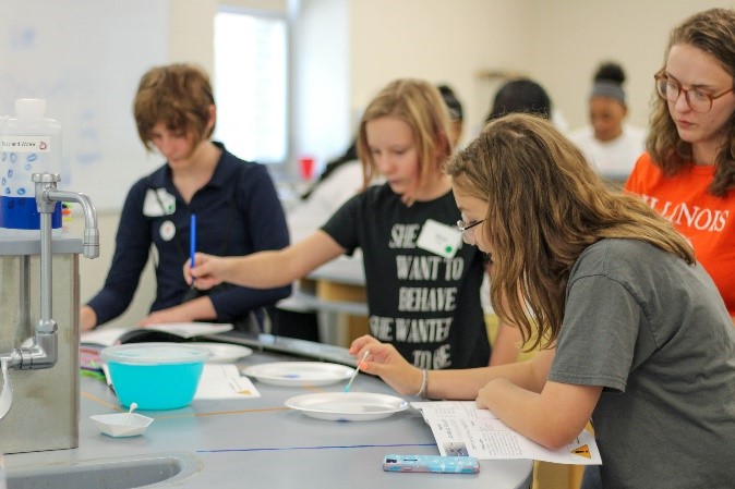 Three girls doing experiments and writing down answers while a volunteer watches.