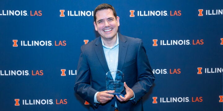 Juan Velásquez holding an LAS alumni achievement award in front of a blue LAS photo backdrop