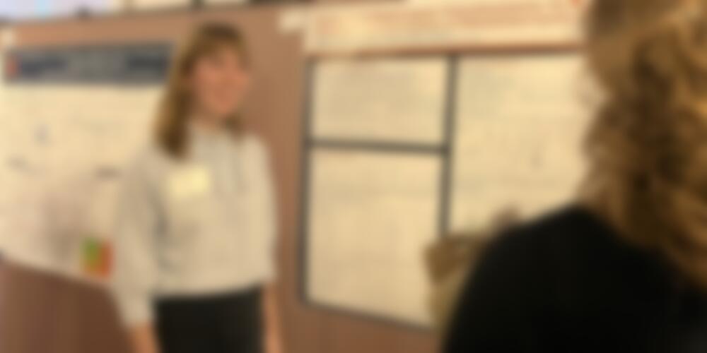 Student stands in front of a research poster on a bulletin board while explaining research to others.