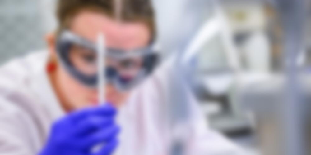 A chemistry student in lab coat and safety goggles and safety gloves is looking closely at a long thin tube she is holding.