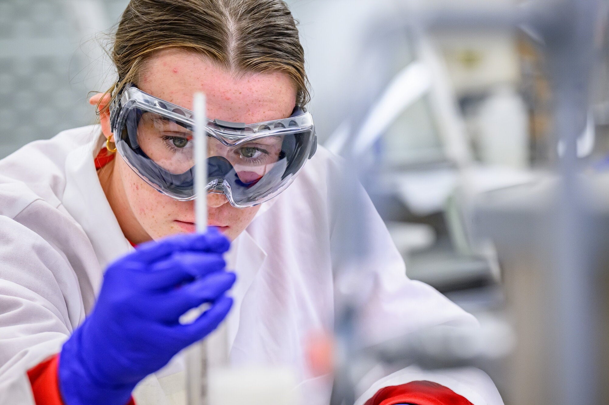 Student in lab coat, goggles and gloves looking at a thin tube