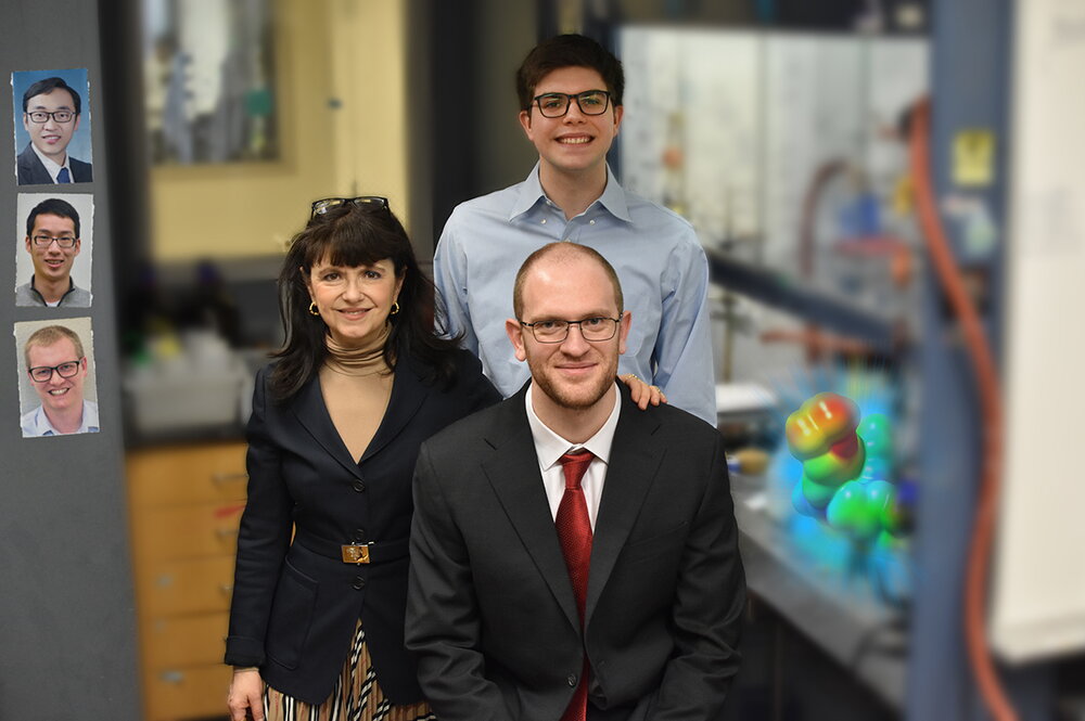 Three researchers stand side by side near equipment in a chemistry lab.