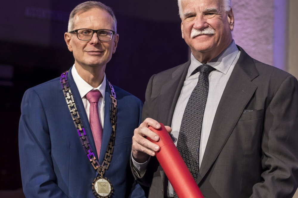 Two men standing side by side in suits and ties with one holding a red certificate tube