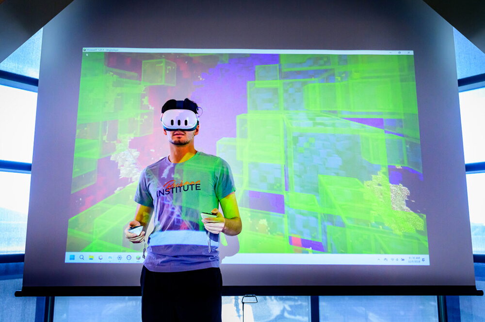 A person wearing a virtual gaming headset and hand controls faces forward in front of a big screen showing the images he is seeing in the headset. 