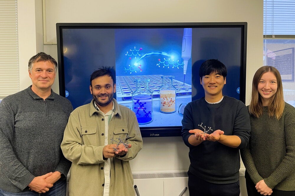 Four people standing in front of a video screen showing a scientific illustration
