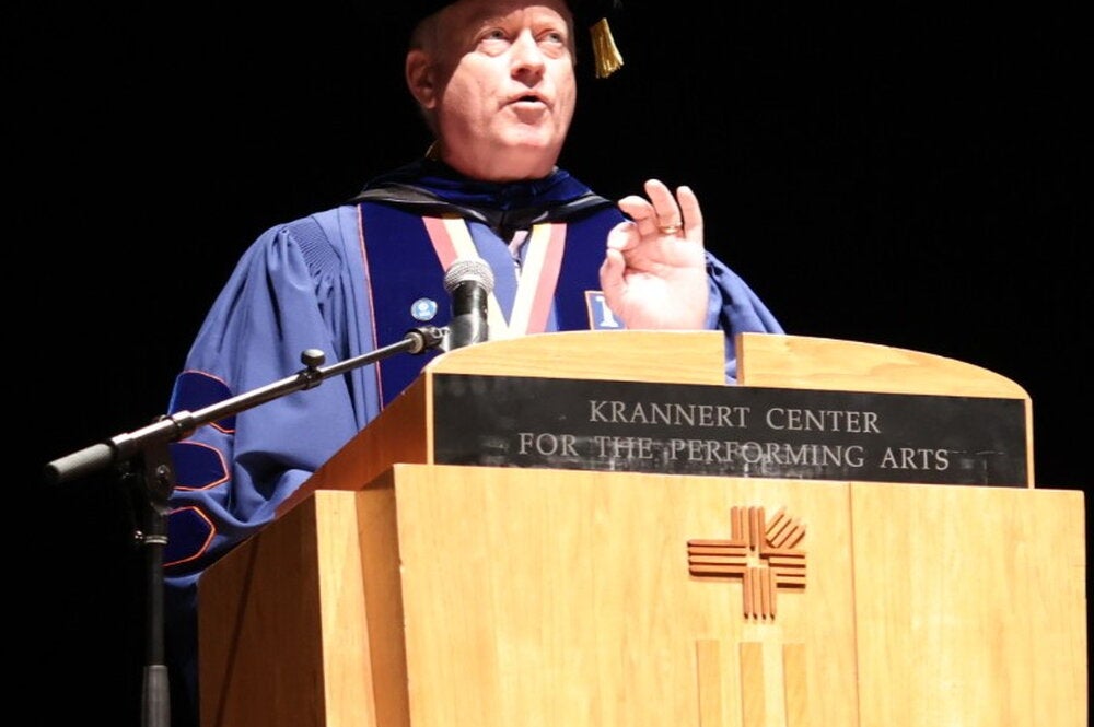 Christopher Cramer in graduation regalia speaking at a podium
