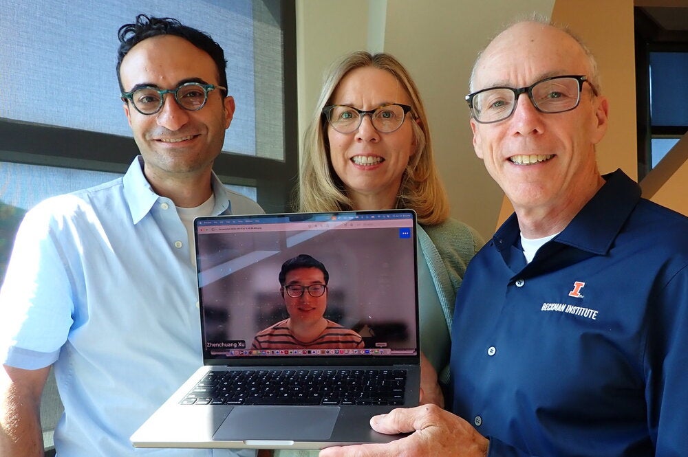 Three people standing side by side holding a laptop with image of a person