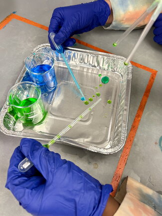 gloved hands working with colored liquids in an aluminum tray