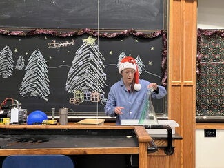 A person in a blue lab coat and holiday hat pouring green liquid into a large flask