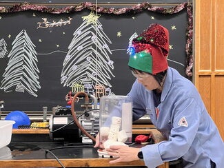 A person in a blue lab coat and holiday hat putting a snowman made of marshmallows under a glass container