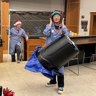 Person in a lab coat and holiday hat with a large plastic garbage can strapped to her 