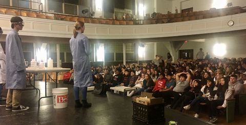Two chemistry students in lab coats stand on a stage in an auditorium full of young students doing chemistry demonstrations.