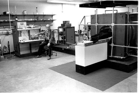 A black and white photo of Paul Lauterbur sitting in a chair near one of the original Big Red MRI machines.