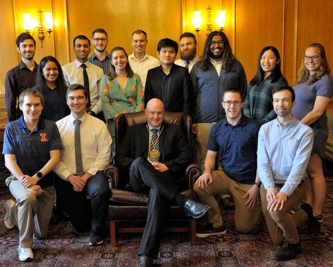 Group photo of Doug Mitchell sitting front row middle in a chair and his research group members kneeling and standing around him in South Lounge of Illini Union.