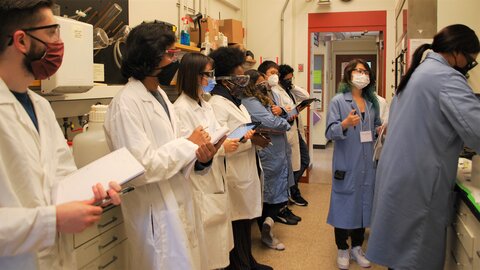 A row of Boot Camp participants in white lab coats and safety gear stand in the lab taking notes as Jeanne N'Diaye in a blue lab coat leads a demonstration.