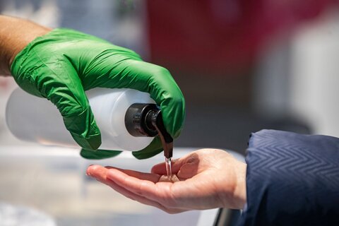 A hand wearing a green rubber glove dispensing hand sanitizer into the bare hand of another person.