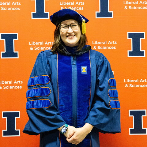 Portrait of Amanda East in graduation regalia standing in front of an orange background with blue Block I