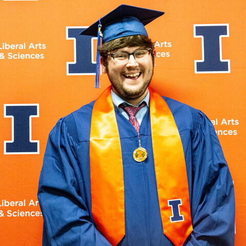 Portrait of Ian Garvey in graduation regalia standing against an orange Block I background