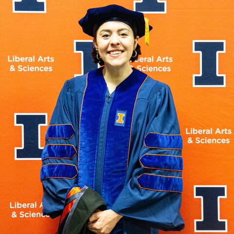 Portrait of Alison Wallum in graduation regalia standing in front of an orange background with blue Block I logos
