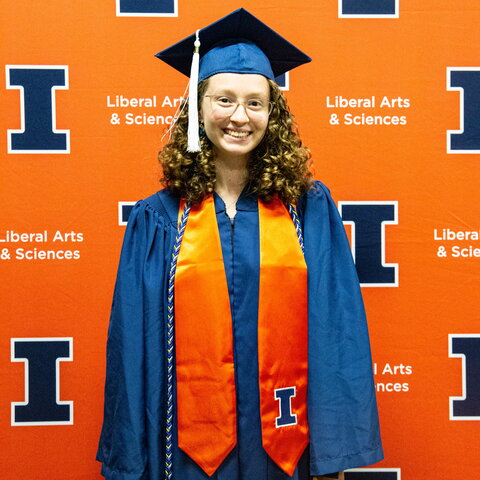 Portrait of Zoe Wachtel in graduation regalia standing in front of an orange background with blue Block I logos