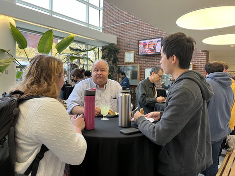 Greg George stands at a table talking with two students during a reception following the lecture.