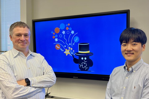 Liviu Mirica and Ju Byeong Chae stand on either side of a television screen mounted on the wall with a photo of a graphic that shows a cartoon figure with a magician's hat and chemical equations. 