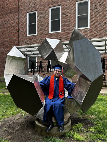 Zach Burke in blue and orange graduation regalia sits in a sculpture outside the CLSL/RAL building on campus.