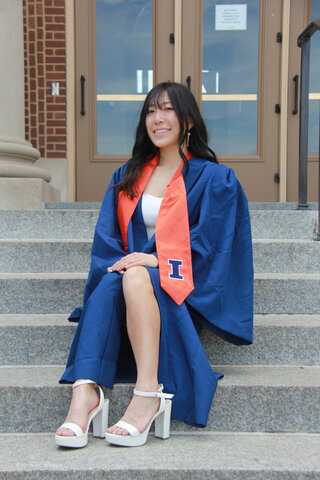 Arisu Oya in orange and blue graduation regalia sitting on steps of campus building.