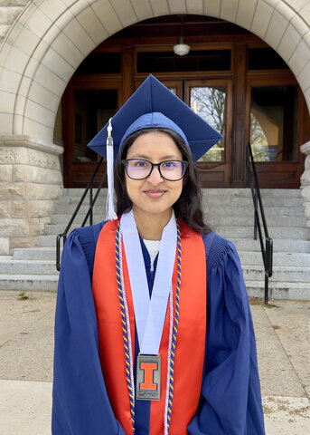 Parmeet Kaur in blue and orange graduation regalia standing in front of Noyes entrance