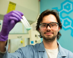 Nathan Gehrke in safety gloves and glasses and lab coat holding a container of liquid.
